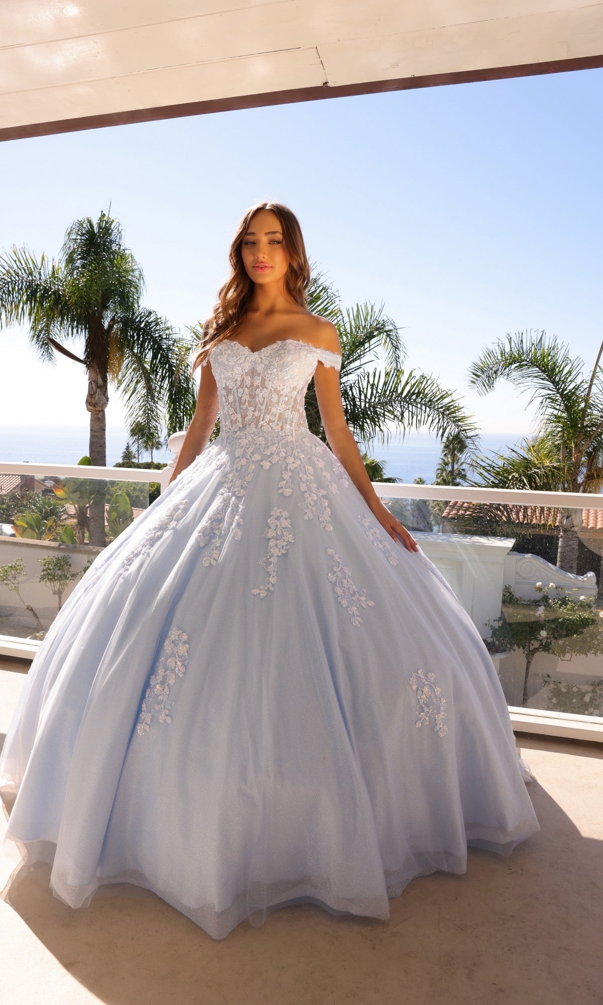 A woman stands on a balcony in the Off-the-Shoulder Lace-Up Long Prom Ball Gown H1352, a light blue embroidered lace dress with floral appliques. Palm trees and a clear sky are seen in the background.